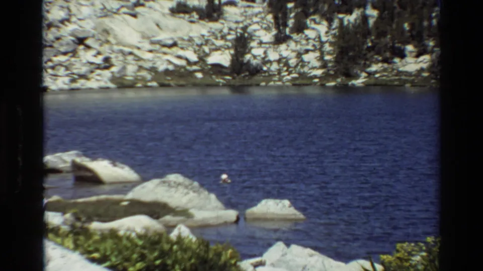 A blue mountain lake fills the frame with pale rocks, a rocky far shore, trees, and a small white object on the water.