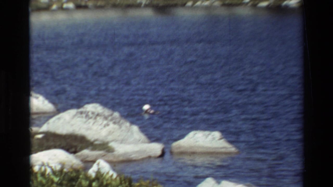 1981 8mm-style frame of a woman swimming near large pale rocks in a blue body of water, with film borders visible.
