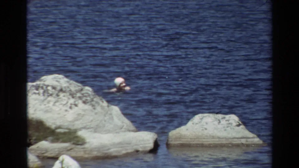 A woman in a white swim cap swims close to pale rocks in blue water, recorded with soft home-movie texture.