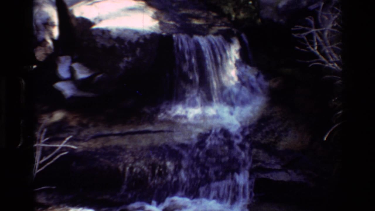 1981 8mm home-movie frame of a small waterfall over dark rocks, with foaming water and a possible partial person at the left edge.