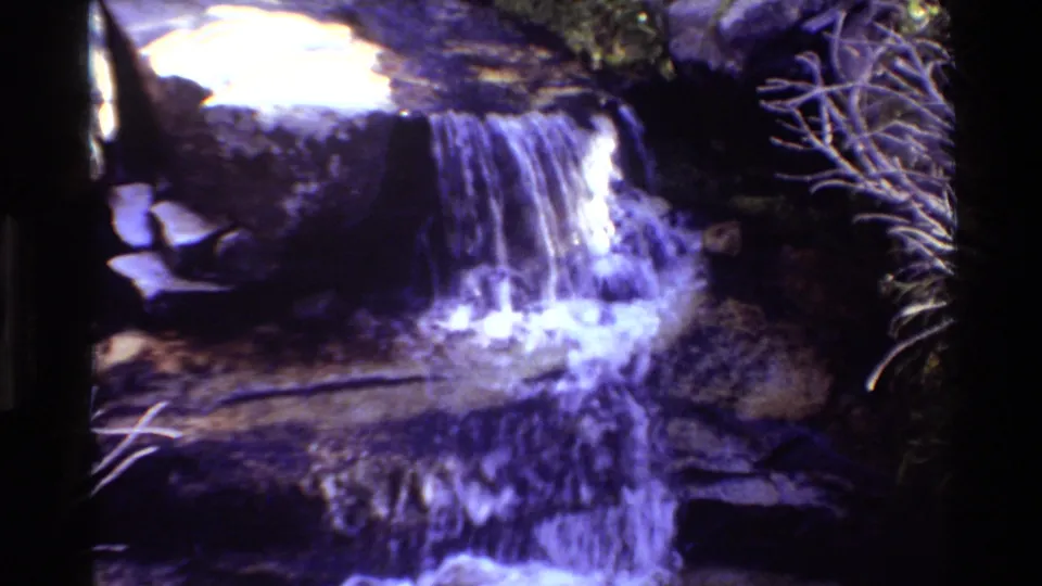 1981 8mm frame showing water falling over a rock shelf with pale branches at the right and strong blue-purple color shift.