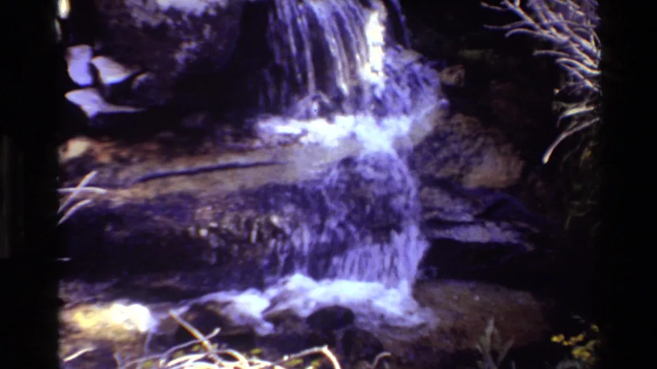 1981 8mm frame of a narrow waterfall running down dark rock into white foam, with foliage visible along the right edge.