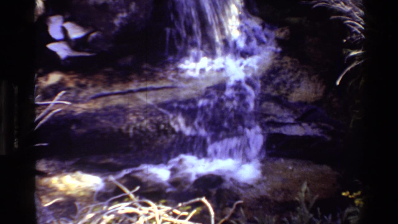 1981 8mm frame of a small waterfall flowing across dark rock ledges, with soft focus and a purple-blue film cast.