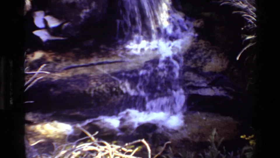 1981 8mm frame of a small waterfall flowing across dark rock ledges, with soft focus and a purple-blue film cast.