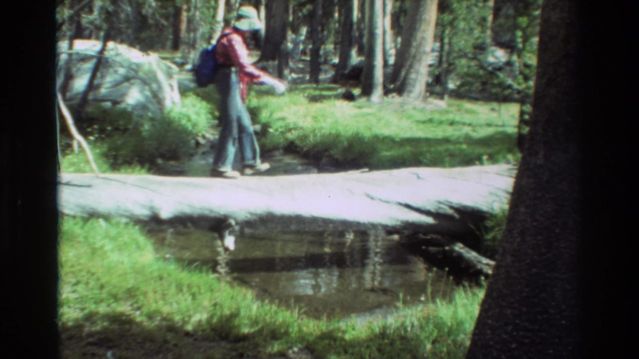 1981 8mm frame of a backpacked hiker in a red shirt and hat crossing a fallen log above a small forest stream.