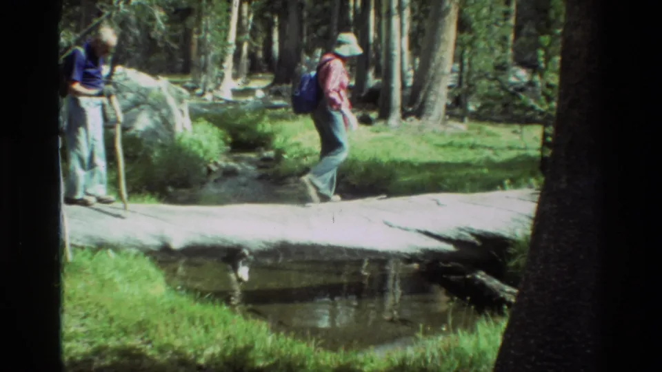 1981 8mm frame showing two backpacked hikers near a fallen-log stream crossing in a wooded setting.