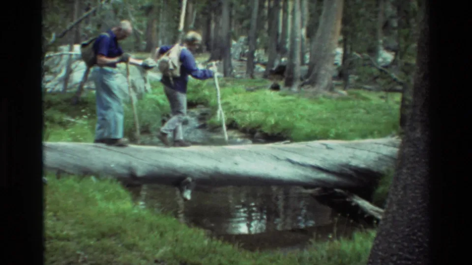 1981 8mm frame showing two backpacked hikers at a forest stream crossing, one holding a walking stick.