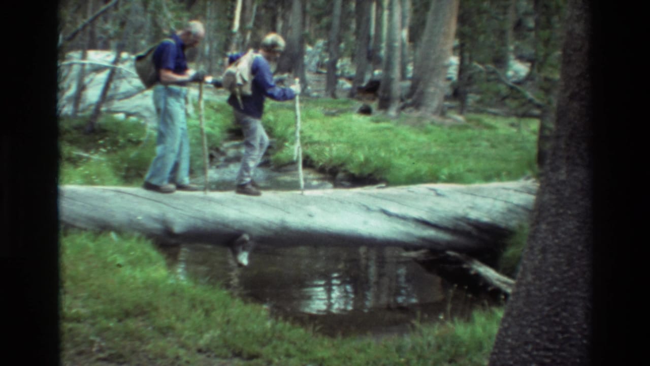 Two older hikers with backpacks and walking sticks pause on a fallen log over a creek in archival 8mm forest footage.