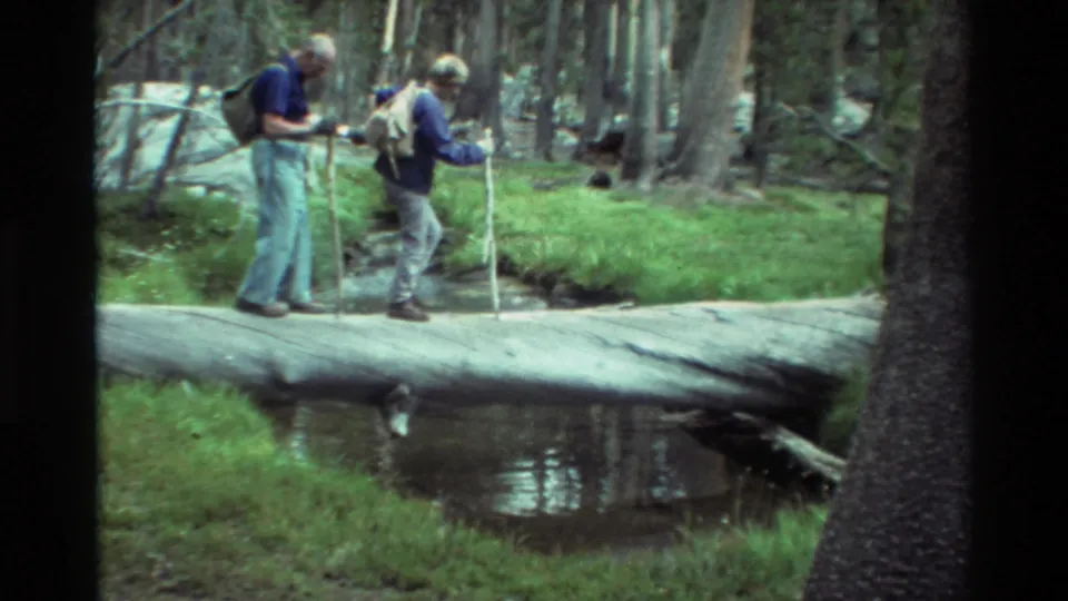 Two older hikers with backpacks and walking sticks pause on a fallen log over a creek in archival 8mm forest footage.