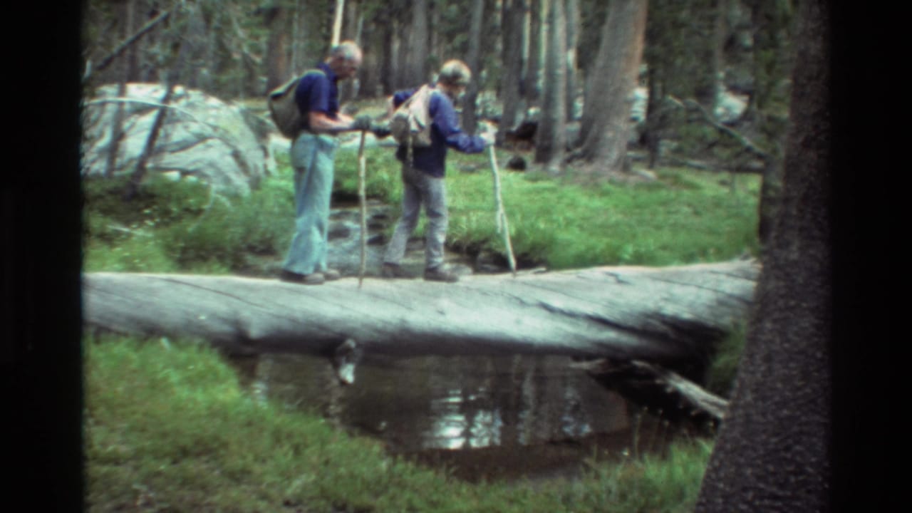 In 1981 home-movie footage, two older hikers use walking sticks while crossing a fallen log over a wooded creek.