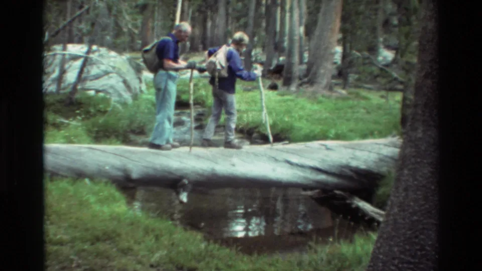 In 1981 home-movie footage, two older hikers use walking sticks while crossing a fallen log over a wooded creek.