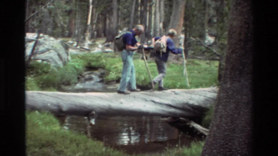 Archival 8mm footage shows two older hikers continuing across a log bridge over a creek in a wooded British Columbia setting.