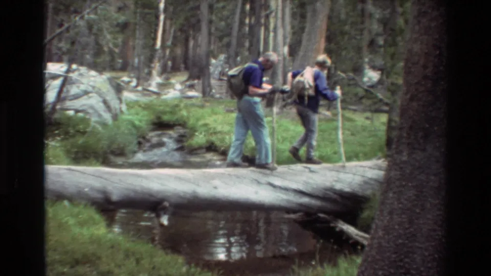 Two adult hikers with backpacks carefully cross a fallen log over water in a dense pine forest, from 1981 8mm home-movie footage.