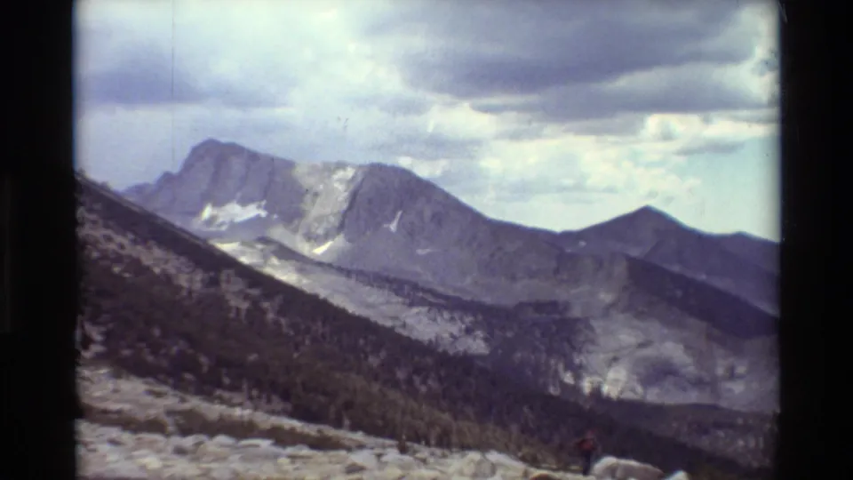 1981 8mm-style wide view of rugged mountain peaks, scattered snow patches, cloudy sky, and dark forested lower slopes.