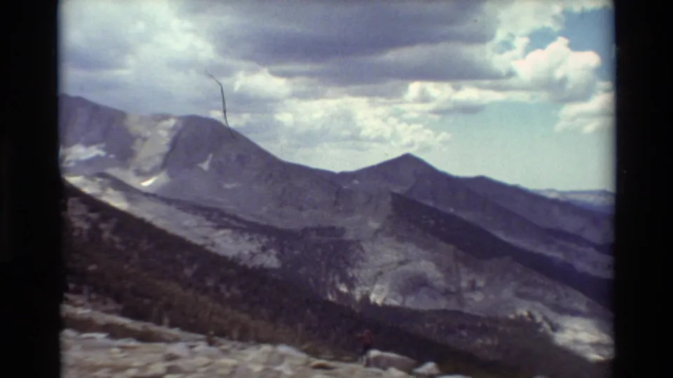 Vintage 8mm frame of a cloudy mountain panorama with a tiny hiker near the lower foreground, providing scale.