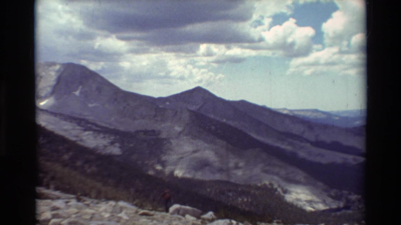 1981 8mm frame of layered mountain ridges, cloudy sky, rocky terrain, and a small hiker visible near the foreground.