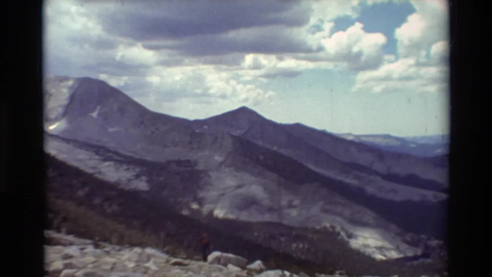 1981 8mm frame of layered mountain ridges, cloudy sky, rocky terrain, and a small hiker visible near the foreground.