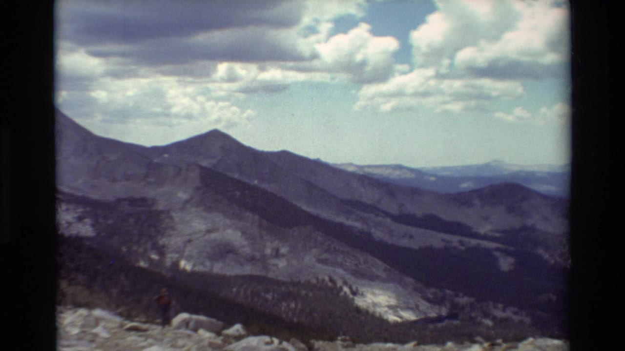 Vintage 8mm mountain panorama with a distant red-shirted hiker standing on rocks beneath bright clouds.