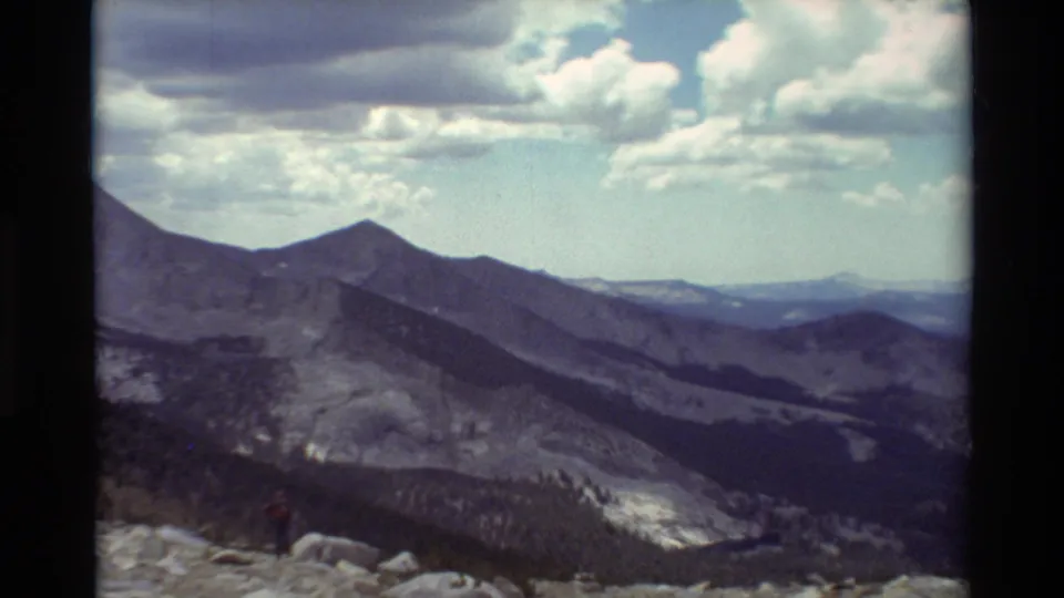 Vintage 8mm mountain panorama with a distant red-shirted hiker standing on rocks beneath bright clouds.