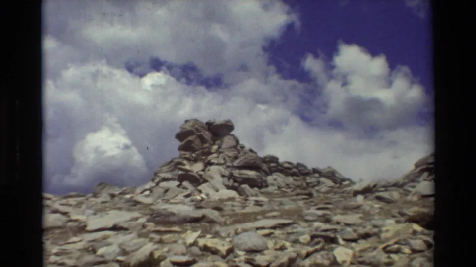 1981 8mm view of a stacked rock cairn on rough alpine stones, framed against a bright cloud-filled sky.