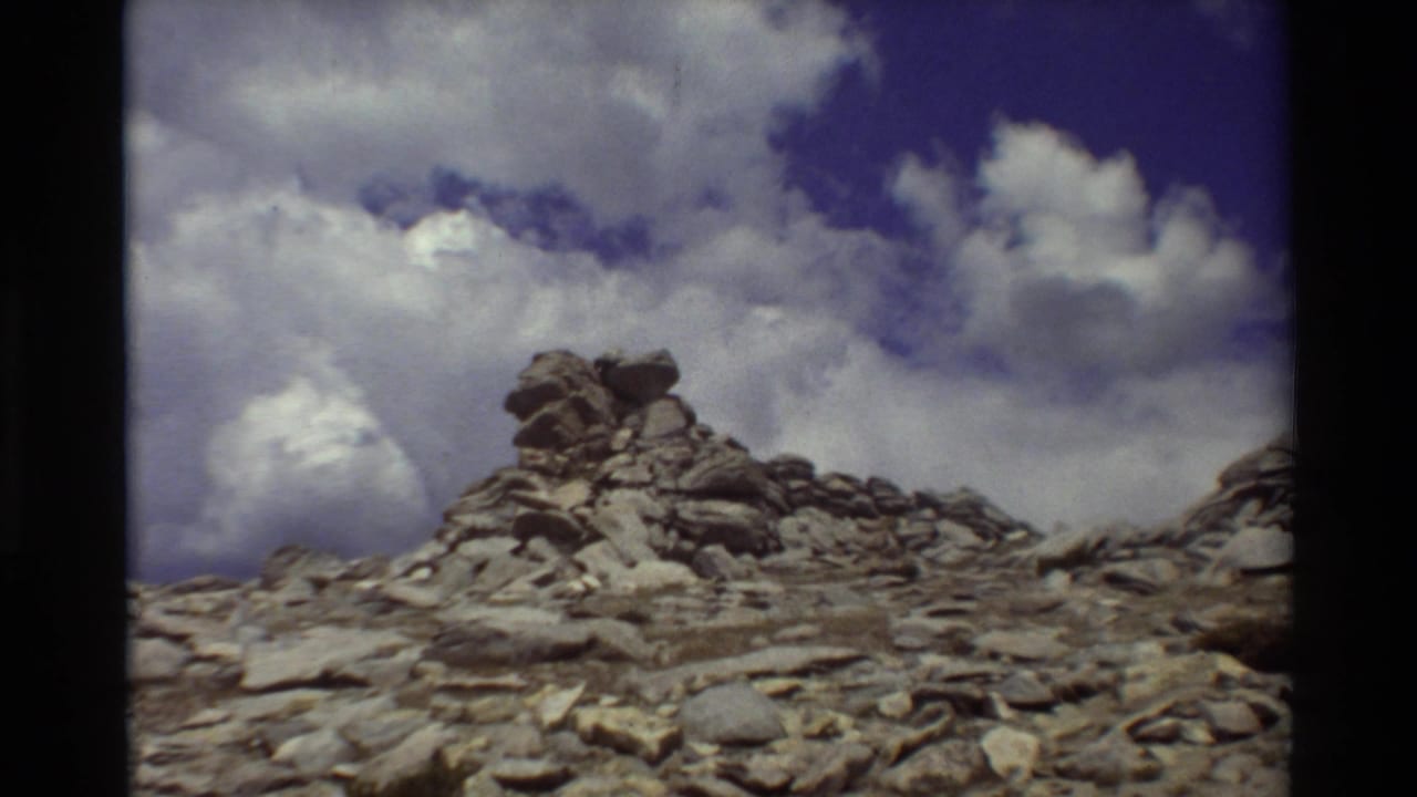 1980s 8mm view of a rocky alpine cairn or outcrop beneath large clouds, with no person clearly visible in this frame.
