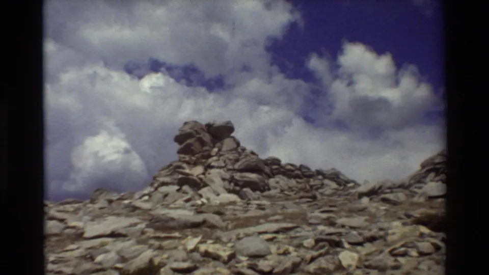 1980s 8mm view of a rocky alpine cairn or outcrop beneath large clouds, with no person clearly visible in this frame.