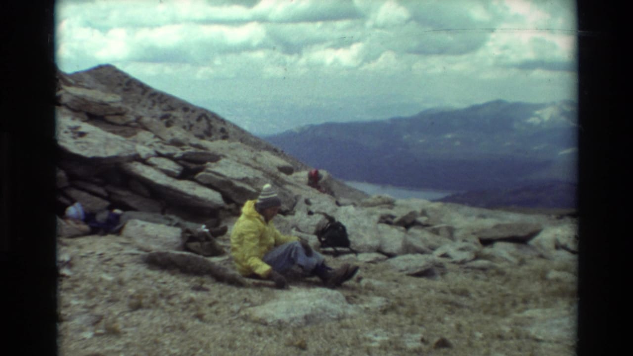 A hiker in a yellow jacket and knit cap sits on a rocky British Columbia mountain slope with distant peaks and a lake in the background.