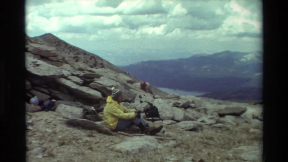 The seated hiker remains on the rocky slope, facing across an alpine view of distant ridges, a lake, and a cloudy sky.