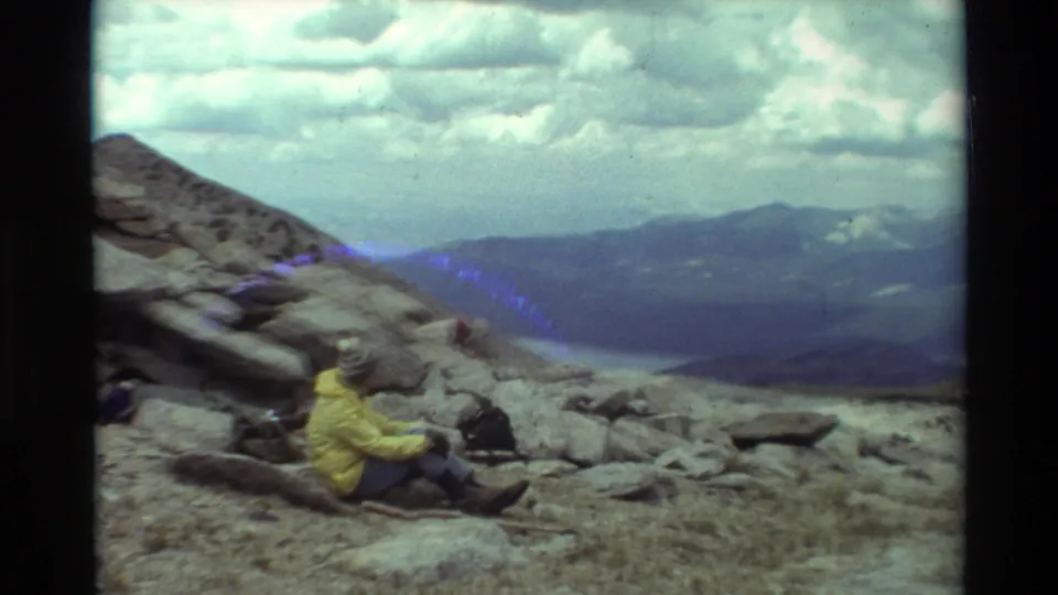 A soft Super 8 frame shows the hiker seated below slanted rocks, with blue light flare, distant lake water, mountain ridges, and clouds.