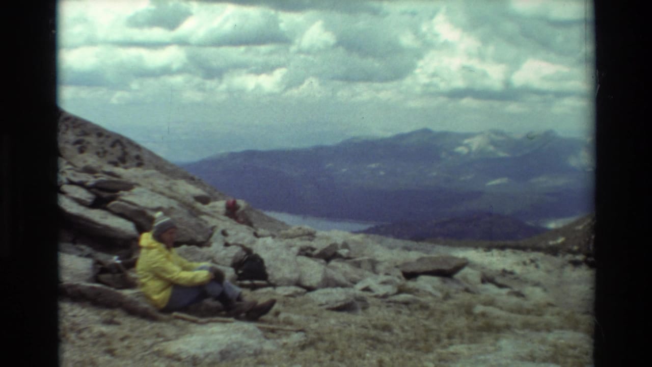 1981 8mm frame of a hiker resting on rocks in a British Columbia mountain landscape, with ridges and heavy clouds beyond.