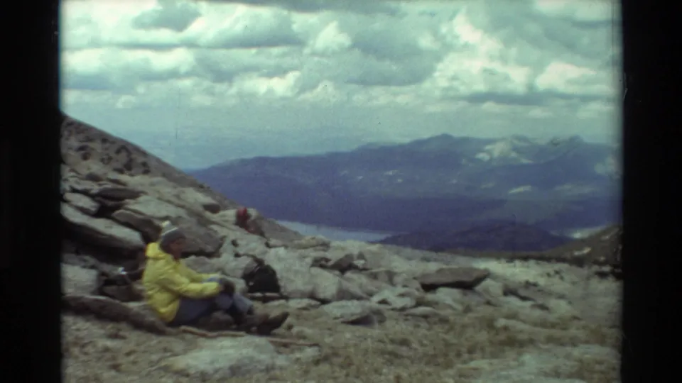 1981 8mm frame of a hiker resting on rocks in a British Columbia mountain landscape, with ridges and heavy clouds beyond.