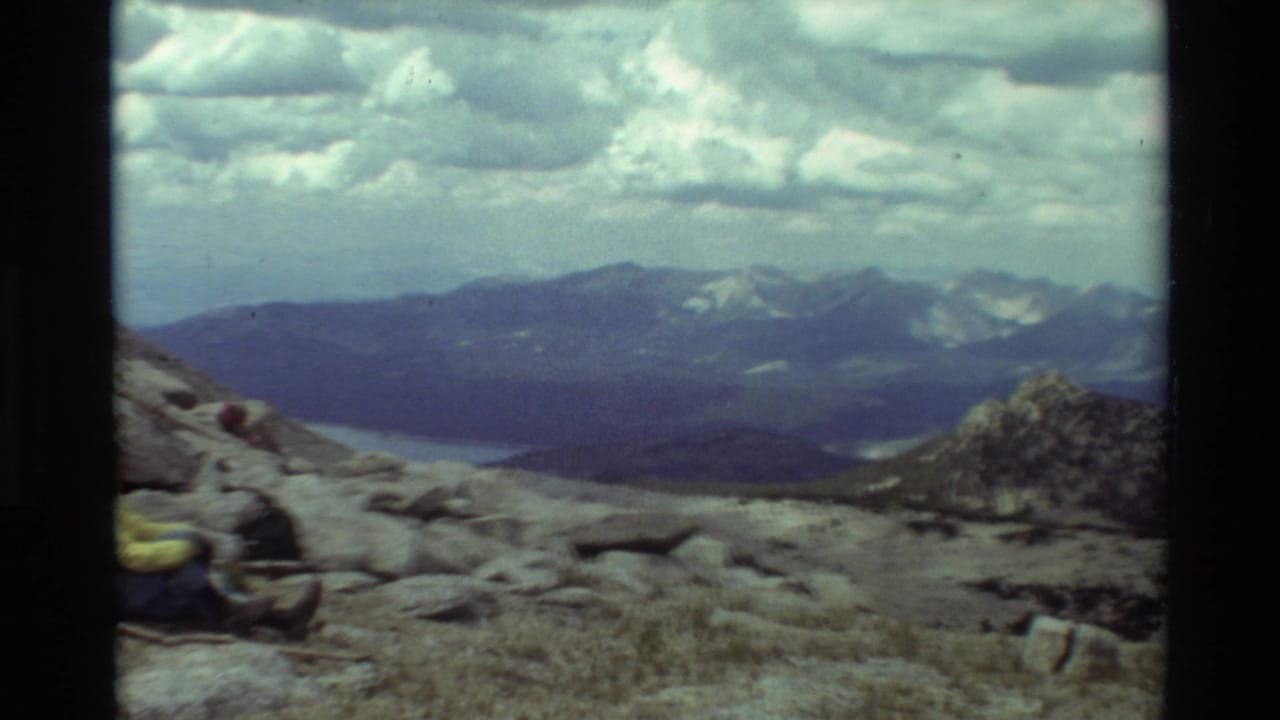 Rocky mountain slope with a partially visible seated hiker and distant ridges under clouds.