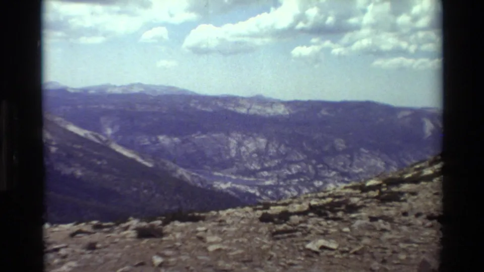 Final 8mm frame looking from a rocky overlook across a broad mountain valley and ridgelines beneath scattered clouds.