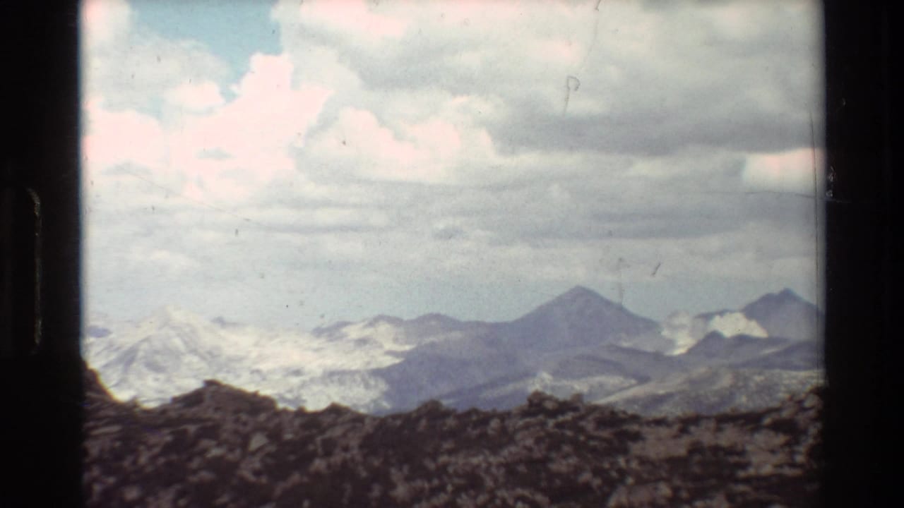 Distant snow-patched mountain range seen beyond a rocky ridge under heavy clouds.