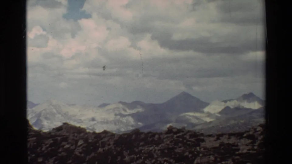 Supported 1981 8mm frame of a cloudy mountain panorama with a low rocky foreground and distant pale snow patches.