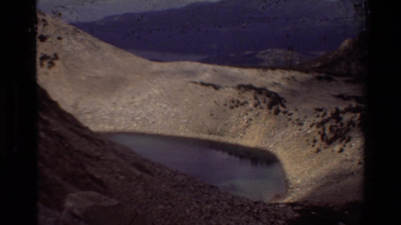 1981 8mm home-movie frame of an alpine lake in a rocky basin with pale slopes and distant mountains, per supplied metadata.