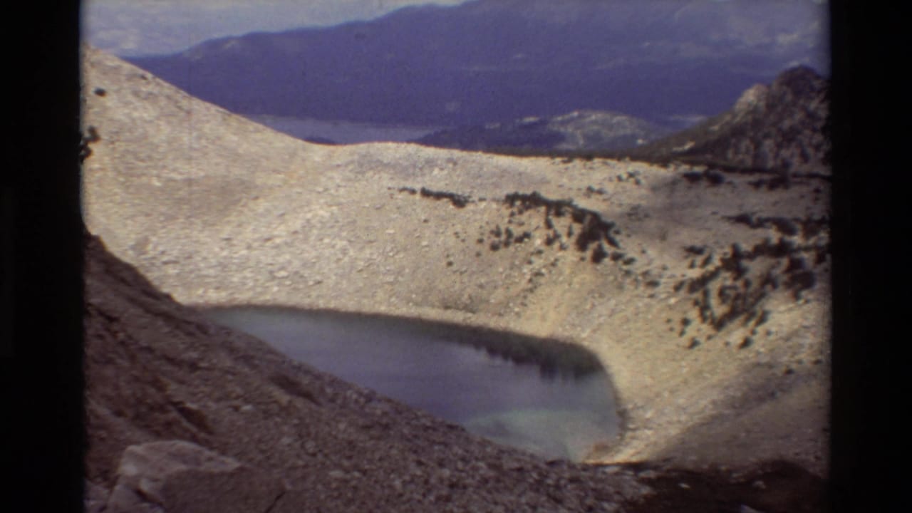 Metadata-listed British Columbia mountain scene with a small alpine lake, snow or scree slope, and distant blue ridges on 8mm film.