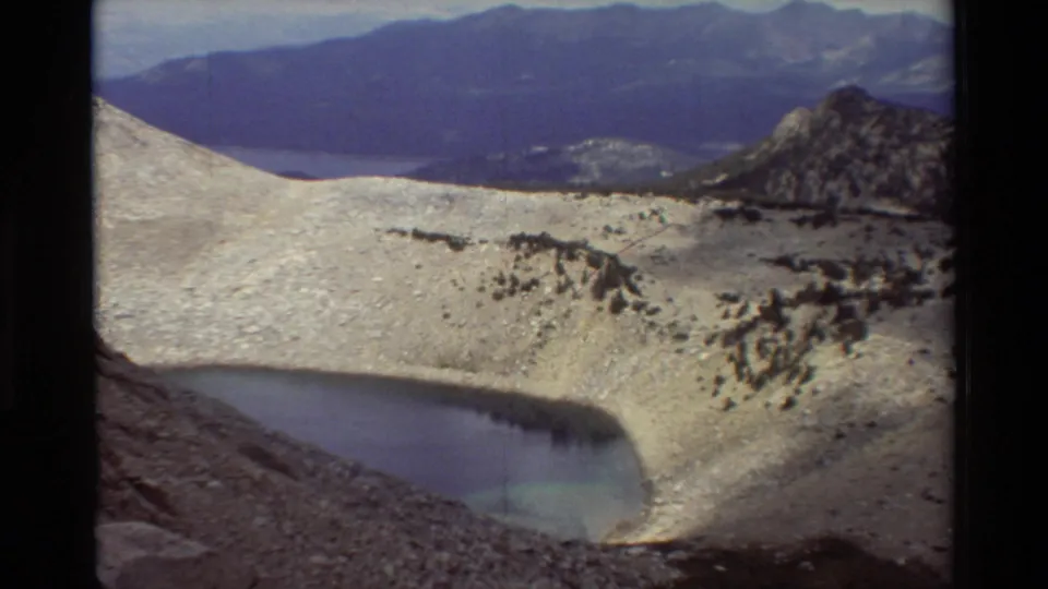 8mm frame of a high mountain basin showing a lake edge, pale rocky or snowy slope, scattered dark rocks, and distant ridgelines.