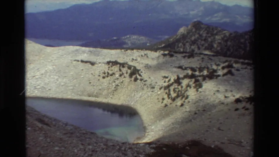 Final 1981 8mm frame shows the alpine lake basin, pale snow or scree, dark rocky outcrops, and layered mountains in the distance.