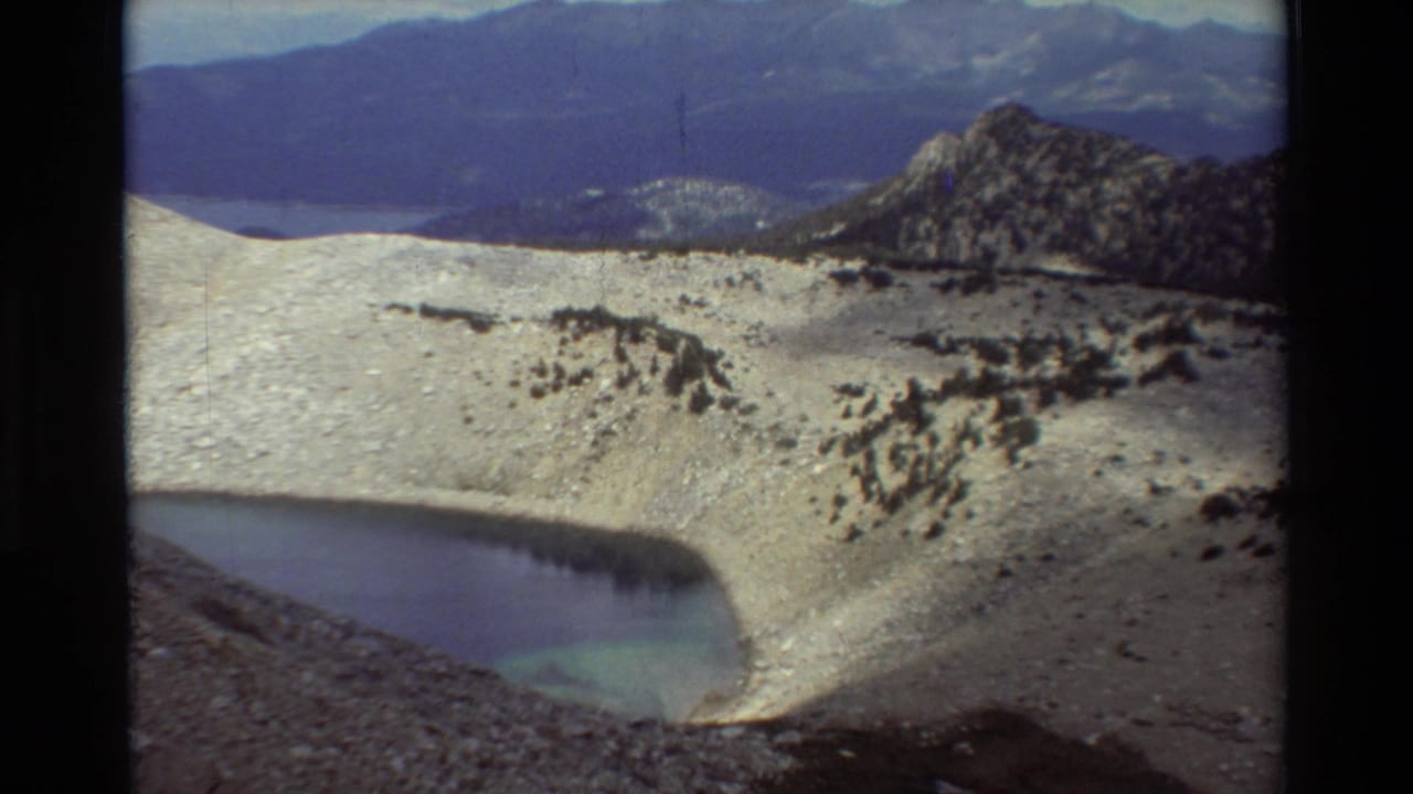 1981 8mm frame of rocky high-country terrain with a small blue-green pond below distant mountain ridges.