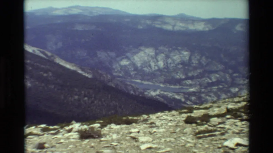 1981 home-movie frame looking across a broad mountain valley from a rocky alpine viewpoint.