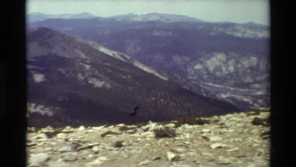 8mm frame of a rocky mountain viewpoint looking toward a distant forested valley and blue ridge horizon.