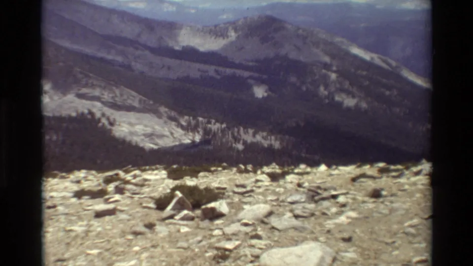 Cataloged 1981 home-movie frame shows rocky foreground ground giving way to a valley and dark mountain slopes.
