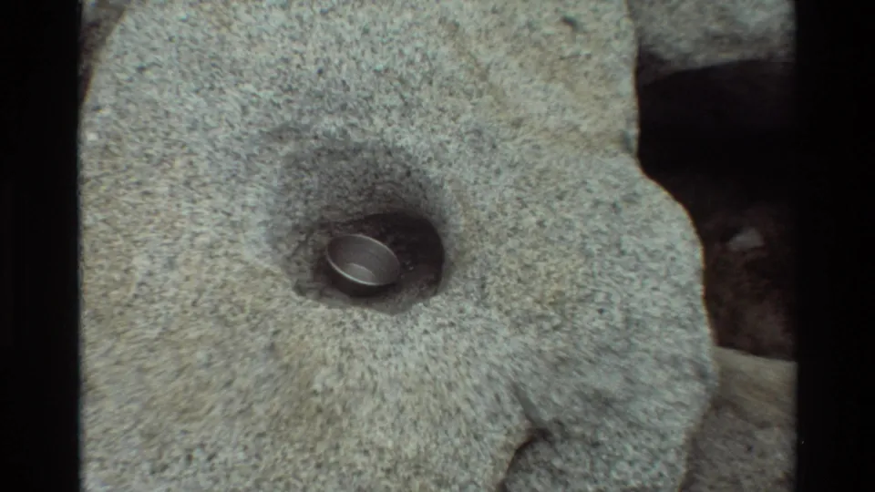 A closer 8mm view shows the reflective metal pot or cup sitting in a shallow hollow within the gray rock.