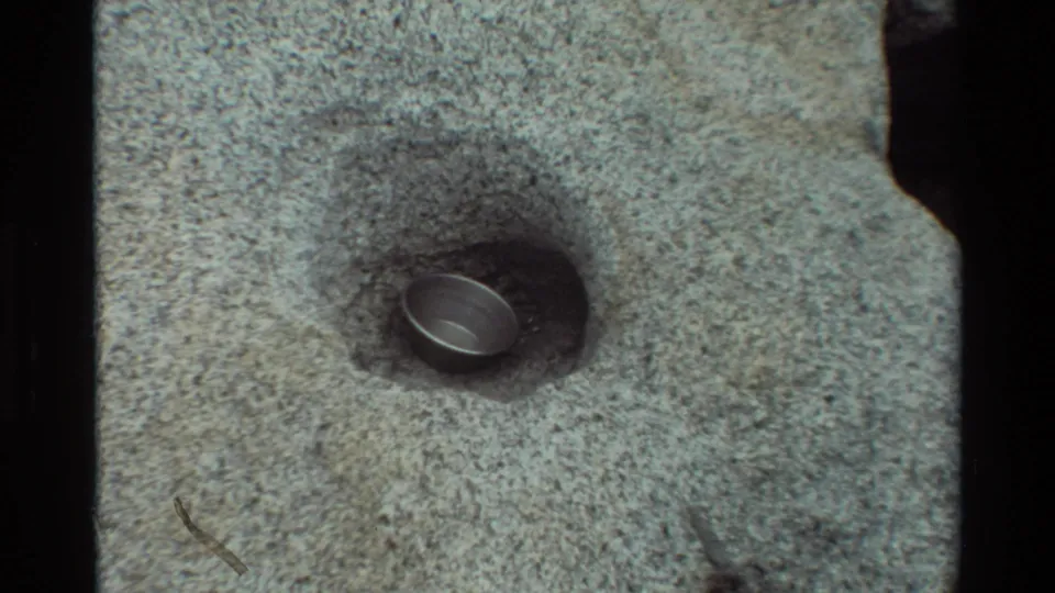 The final frame keeps the small metal pot or cup centered in a shallow rock hole, with rough gray stone filling the view.