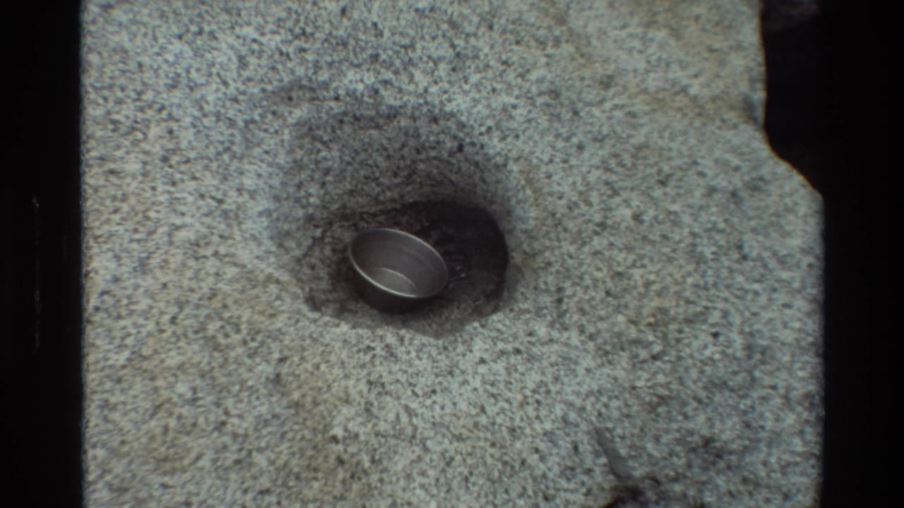 1981 8mm frame from British Columbia metadata showing a small metal object set into a shallow hollow in coarse gray rock.