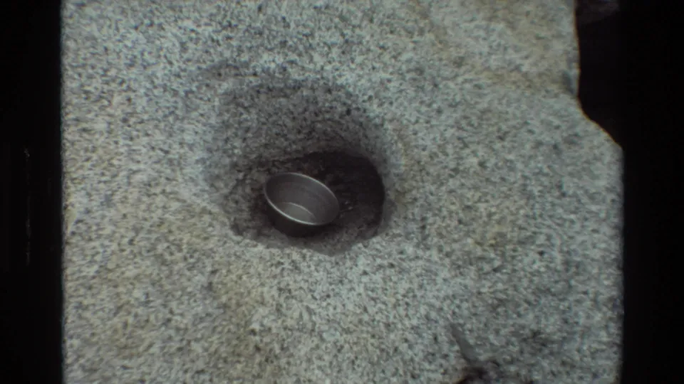 1981 8mm frame from British Columbia metadata showing a small metal object set into a shallow hollow in coarse gray rock.