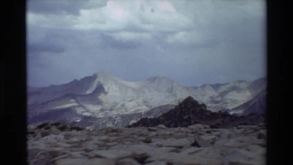 Vintage 8mm view of a rocky alpine foreground with distant snow-dusted mountains and overcast sky, location per metadata.