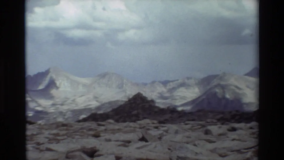 Wide 8mm mountain landscape showing a rocky field, dark midground ridge, pale distant peaks, and overcast sky.
