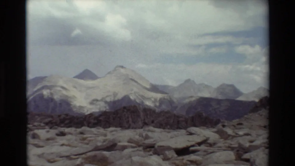 1981 home-movie frame showing distant snowy alpine peaks, dark ridgelines, and a rocky foreground under a bright cloudy sky.